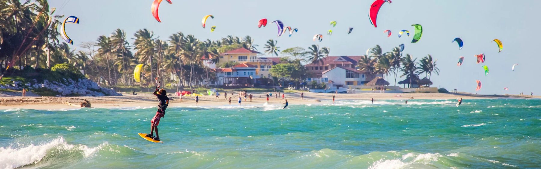 Kitesurfers on the water at Cabarete beneath a sky full of colourful kites, with palm trees and beachfront buildings behind