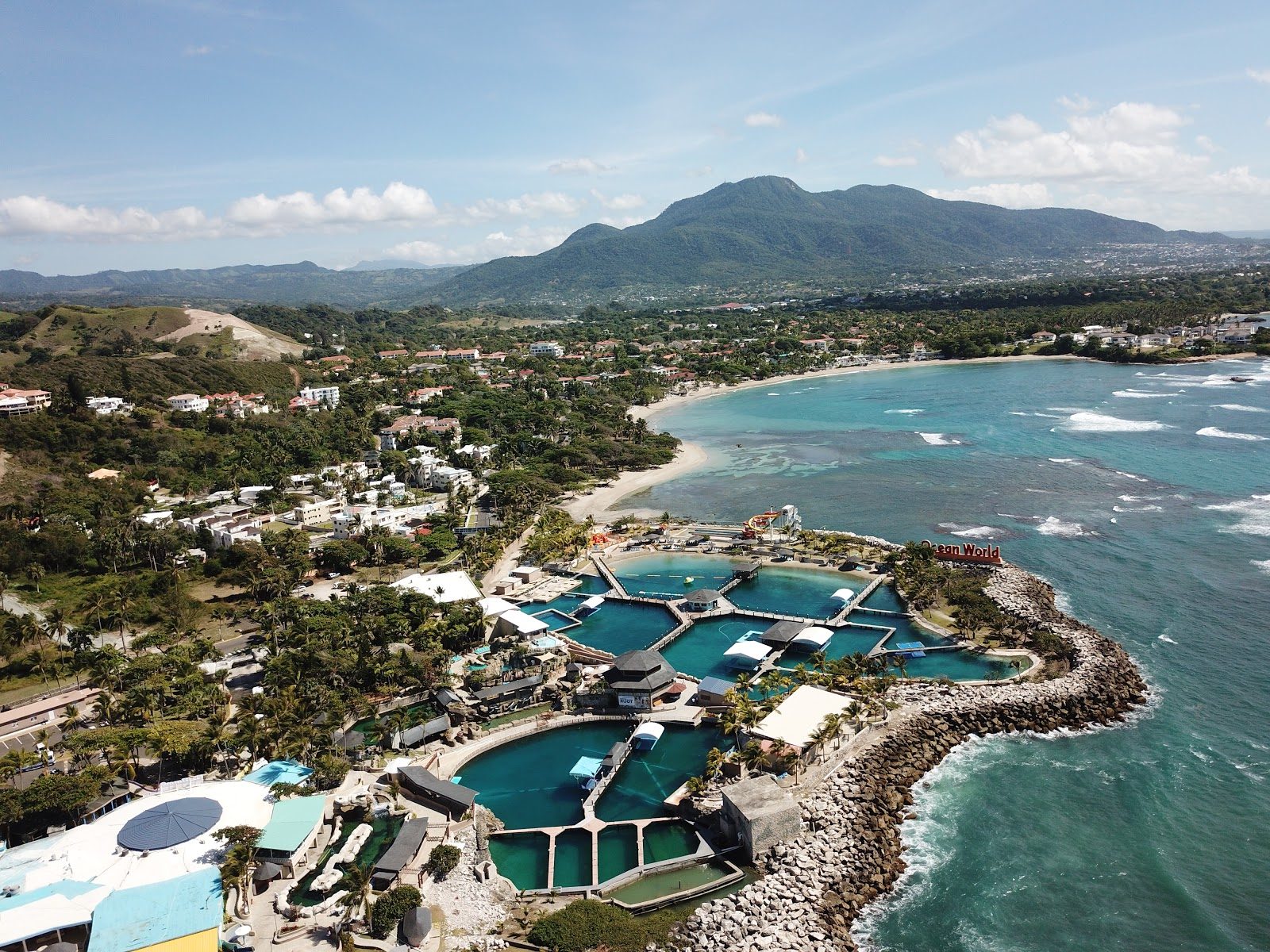 Aerial view of Cofresí: the Ocean World Adventure Park lagoons on the rocky point, the cove beach and resort strip behind, and Pico Isabel de Torres rising inland