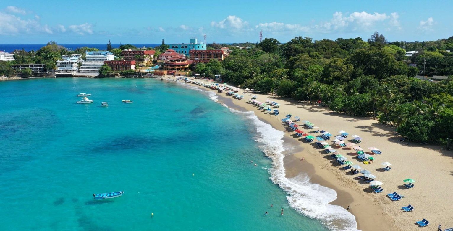 Aerial view of Playa Sosúa: the turquoise bay, rows of beach umbrellas along the sand and the town's hotels on the headland above